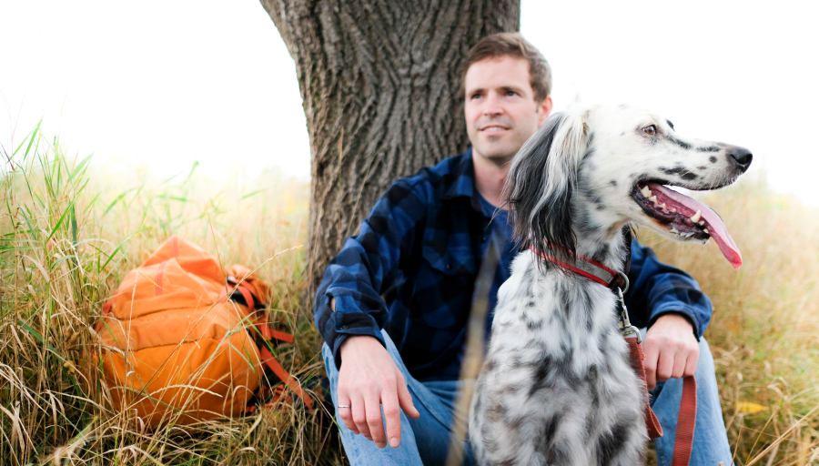 De man zat voor een boom met zijn oranje rugzak op het gras en zijn hond zat met verlof voor hem